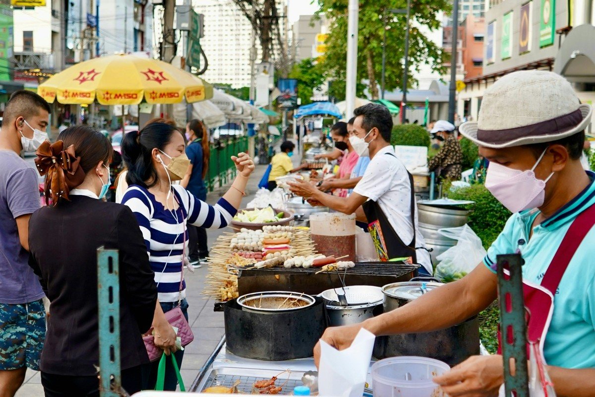 Étal de rue à Bangkok où un vendeur fait griller des brochettes de viande en plein jour