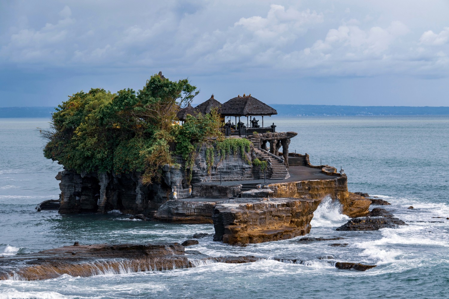 Le temple de Tanah Lot sur son îlot rocheux, entouré par les vagues à marée haute, sous un ciel couvert
